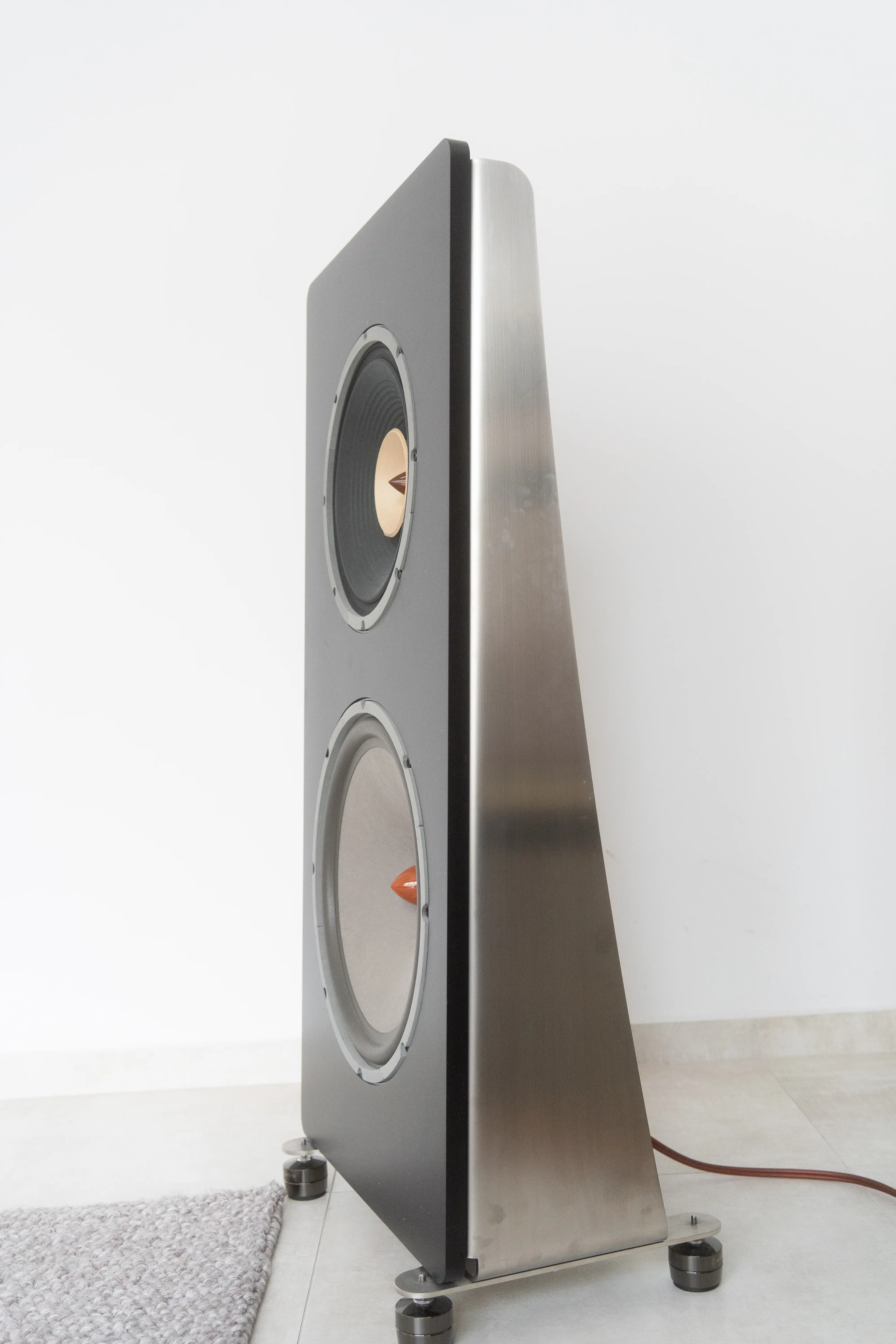 House of Stain 4k loudspeaker, 3/4 angle: dark baffle with two LiiAudio drivers (F-12 above the W-15), wrapped on one side by a curving brushed-stainless U-frame. The open back is visible. Floor-standing on metal feet.