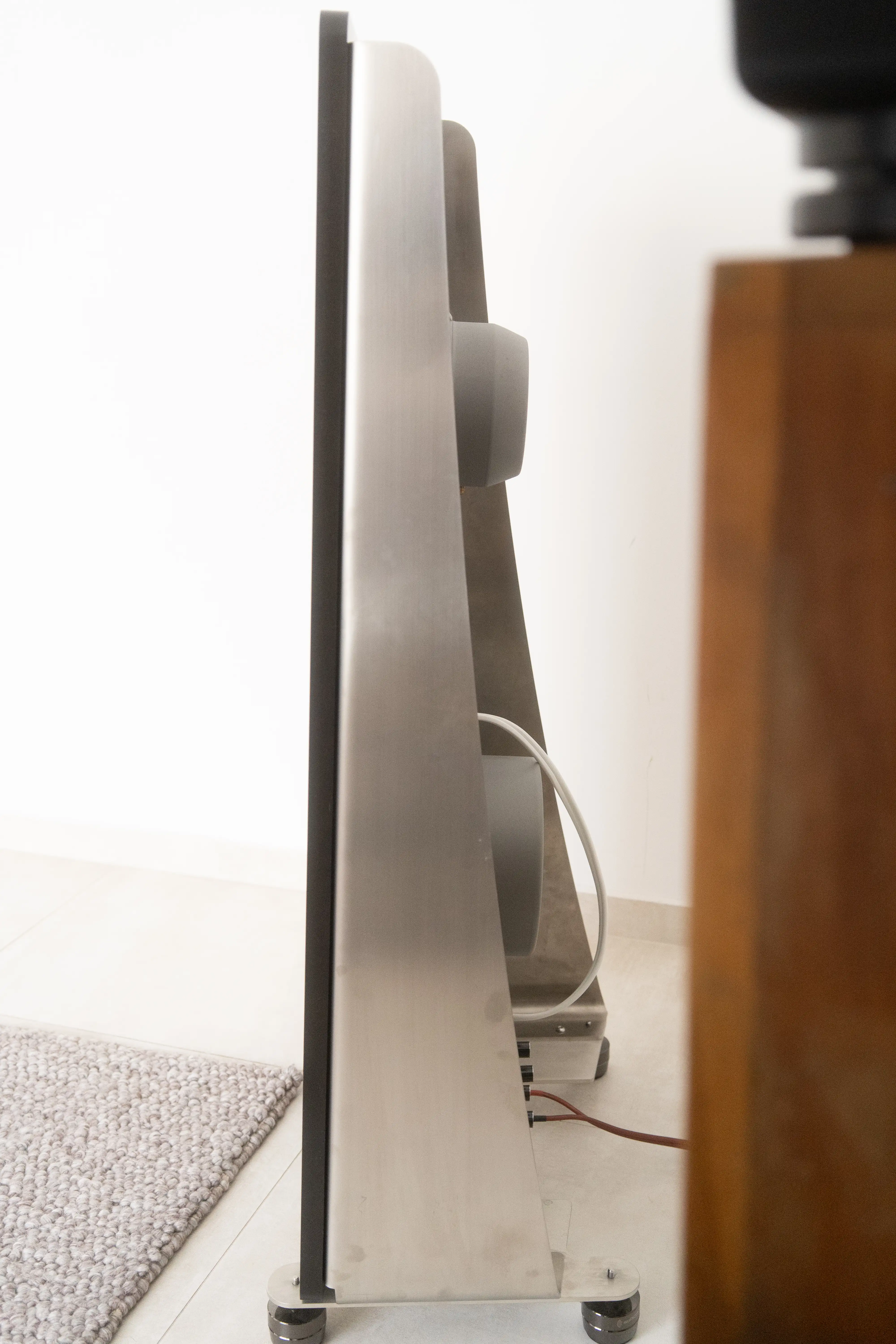House of Stain 4k seen in profile — the asymmetric stainless-steel U-frame curves from baffle to back, the open rear lets the drivers radiate behind. Mounted on steel feet.